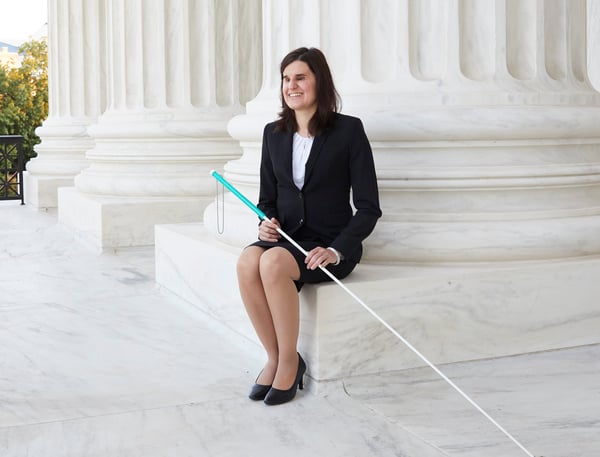 Laura Wolk is wearing a black skirt suit with a white blouse and is sitting outside the Supreme Court building, holding her cane.