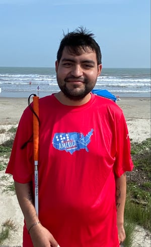 Ruben Rincon wearing a red Braille Across America t-shirt is standing with his white cane in hand at a beachy trail.