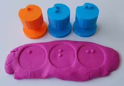 A consonant and two vowel stampers in bright blue and orange above a stamped bit of play-dough that shows the braille letters that were pressed into it.