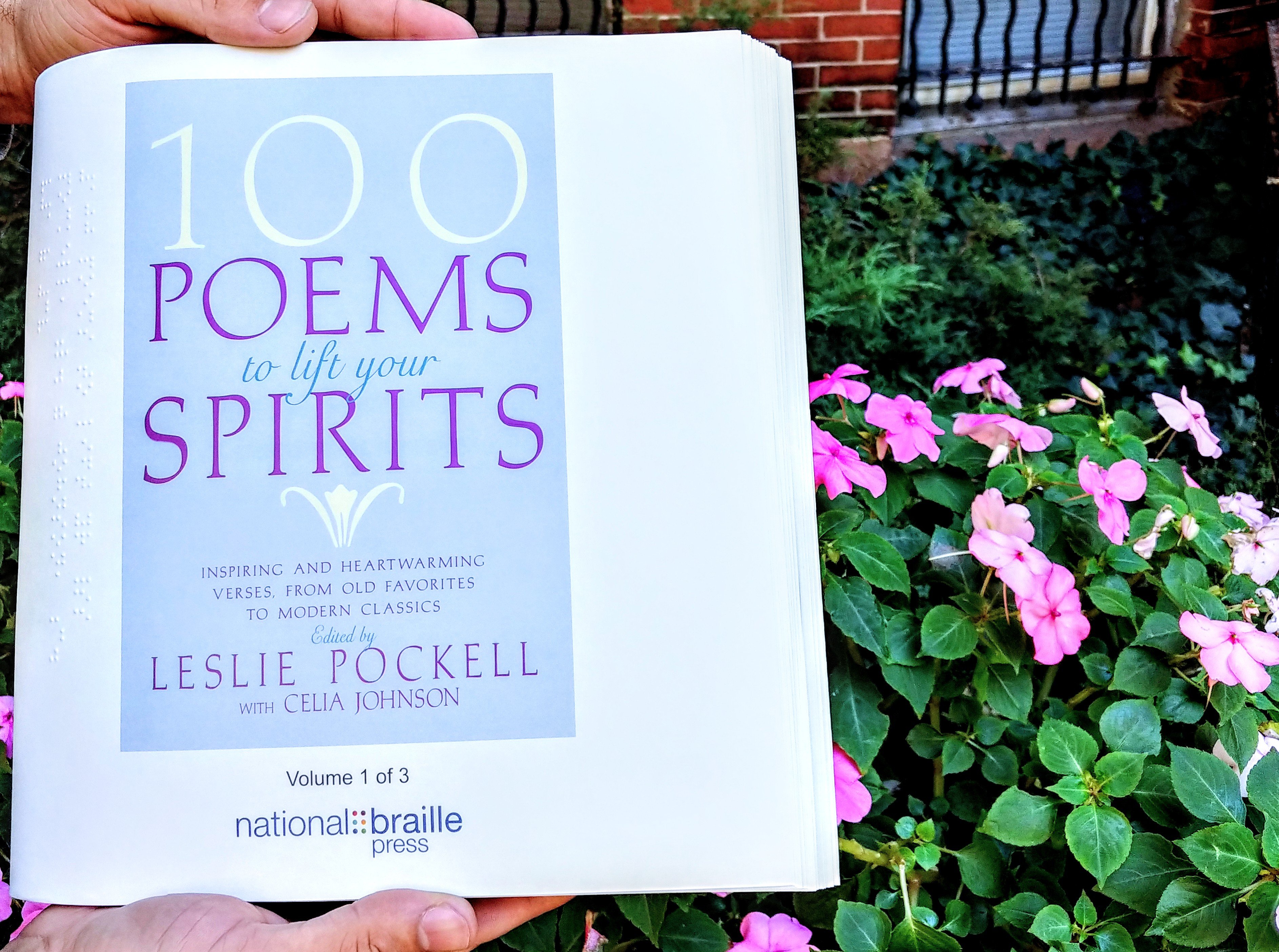 100 Poems to lift your spirit book cover held by someone stading next to some pink flowers.