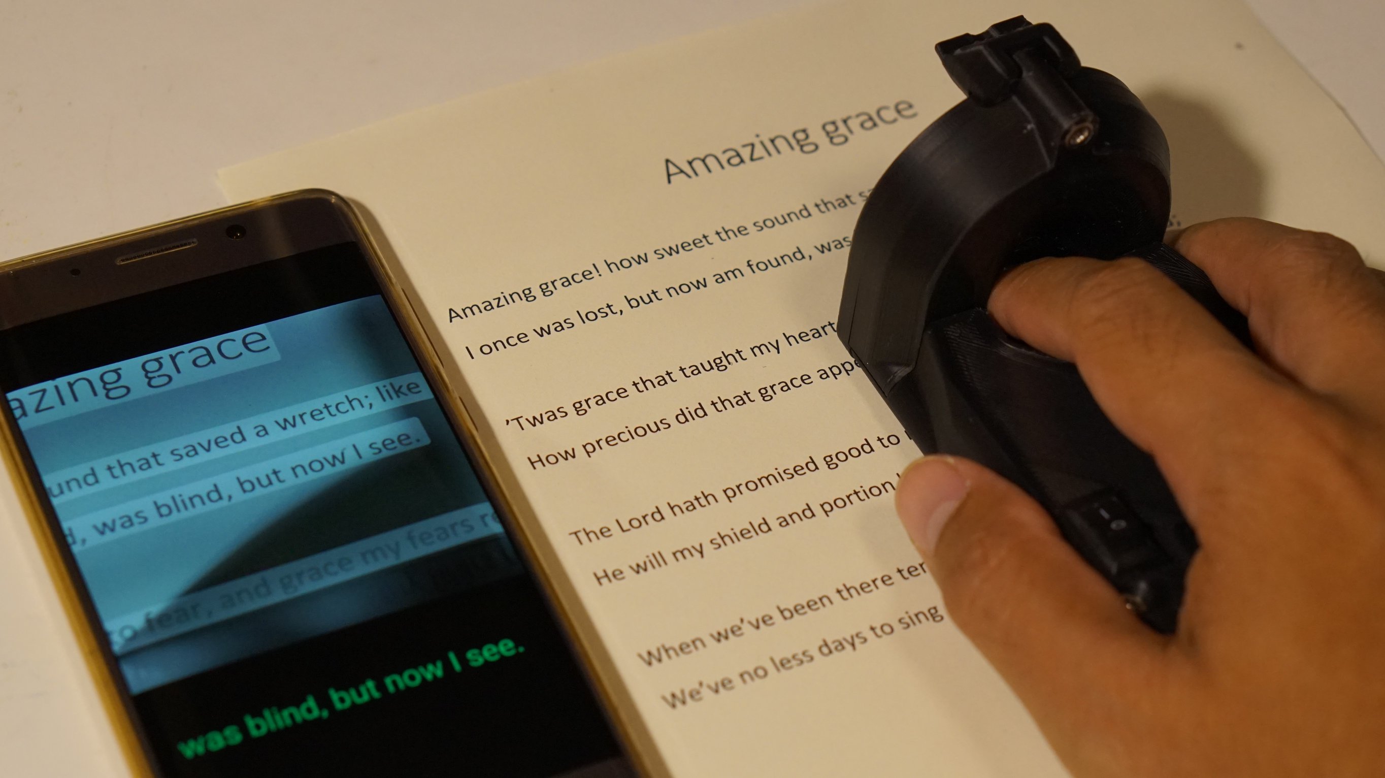 The ReadRing in action. Its scanning the text of Amazing Grace on paper that's showing up on a smartphone that's triggering the rotary braille display in the ReadRing.